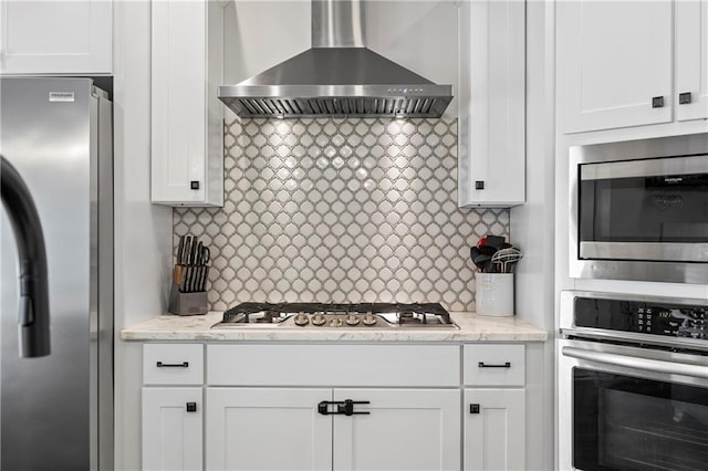 kitchen featuring light stone counters, white cabinets, appliances with stainless steel finishes, wall chimney range hood, and decorative backsplash