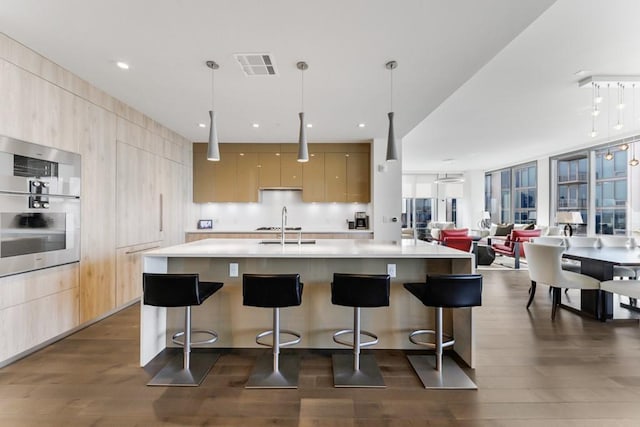 kitchen with light brown cabinetry, dark hardwood / wood-style flooring, a kitchen island with sink, hanging light fixtures, and a breakfast bar area