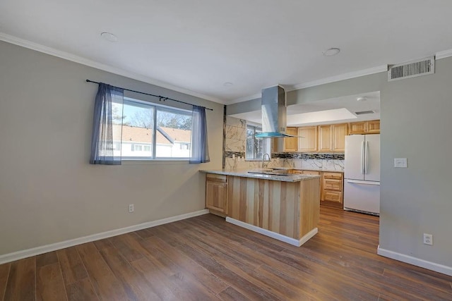 kitchen featuring kitchen peninsula, island range hood, white fridge, backsplash, and light brown cabinets