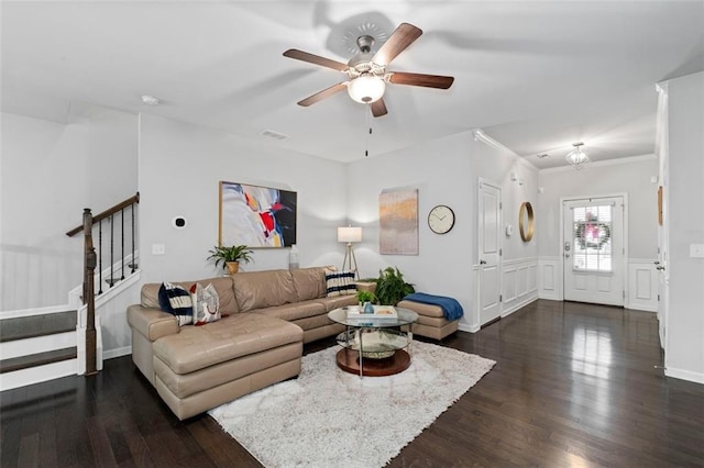 living room with dark wood-type flooring, a ceiling fan, stairs, a wainscoted wall, and crown molding