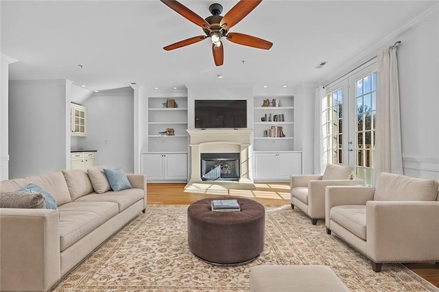 living area with a glass covered fireplace, built in features, a ceiling fan, wood finished floors, and visible vents