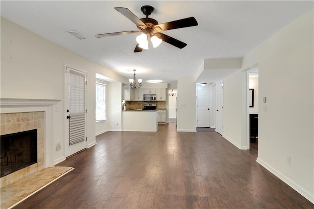 unfurnished living room with a chandelier, a ceiling fan, a tiled fireplace, dark wood-type flooring, and baseboards