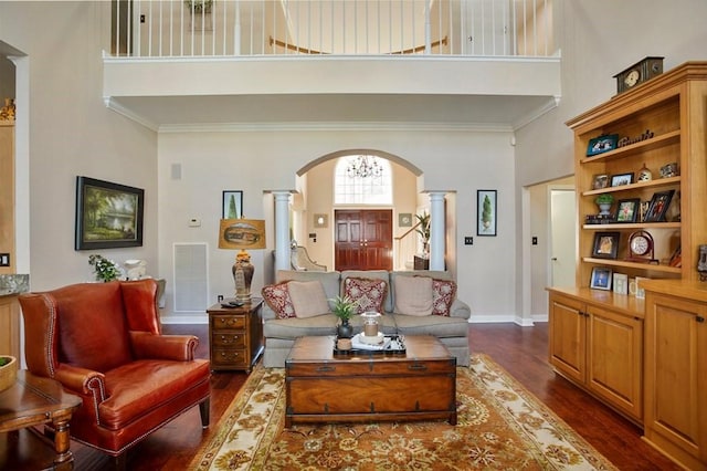 living room featuring a towering ceiling, decorative columns, arched walkways, dark wood finished floors, and crown molding