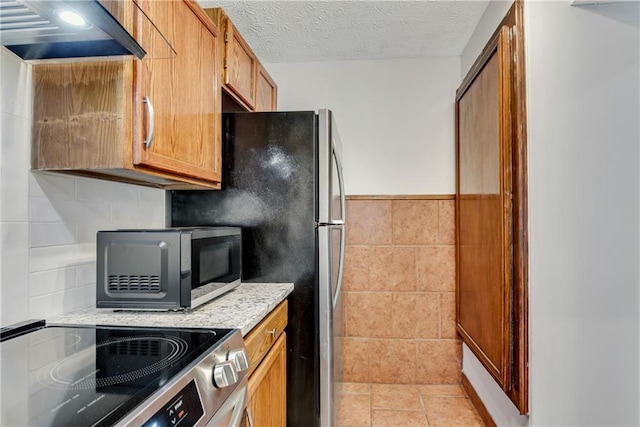 kitchen featuring a textured ceiling, tile walls, appliances with stainless steel finishes, brown cabinets, and custom range hood