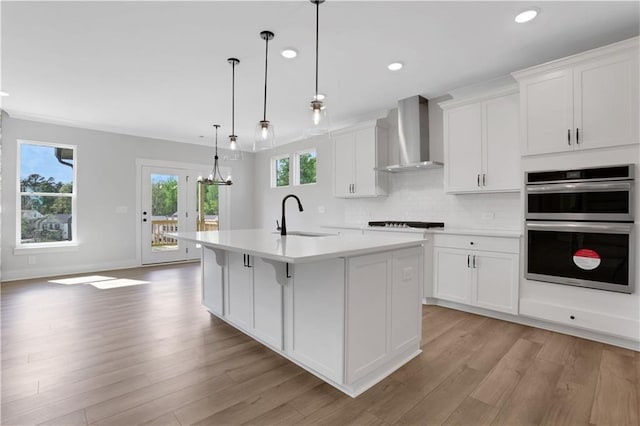 kitchen featuring white cabinets, wall chimney exhaust hood, a sink, an island with sink, and double oven