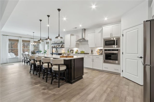 kitchen with wall chimney exhaust hood, an island with sink, pendant lighting, stainless steel appliances, and white cabinets