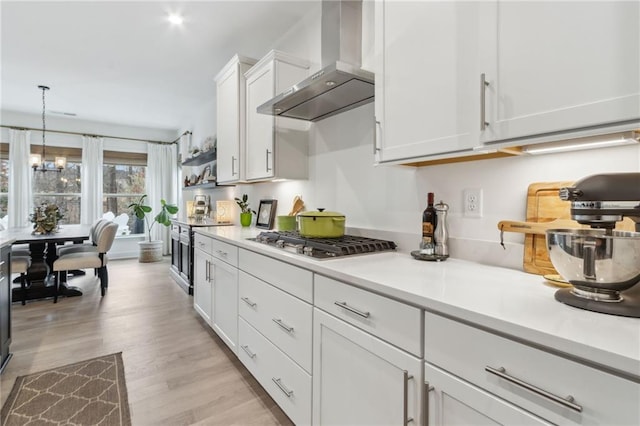 kitchen with wall chimney exhaust hood, white cabinetry, hanging light fixtures, light wood-type flooring, and stainless steel gas stovetop