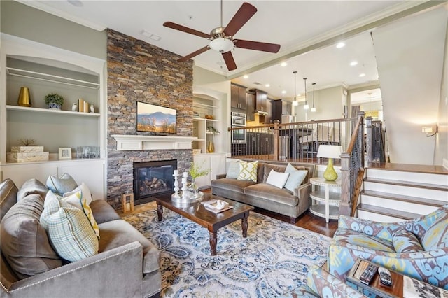 living room with crown molding, ceiling fan, hardwood / wood-style floors, built in shelves, and a stone fireplace