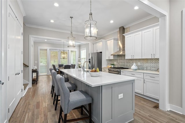 kitchen with wall chimney exhaust hood, appliances with stainless steel finishes, an island with sink, tasteful backsplash, and ornamental molding