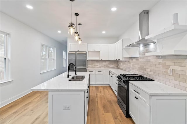 kitchen with stainless steel appliances, wall chimney range hood, a center island with sink, white cabinetry, and hanging light fixtures