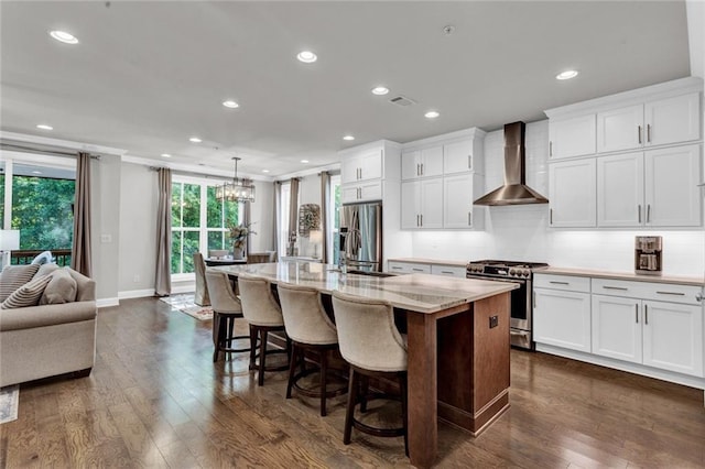 kitchen with an island with sink, dark hardwood / wood-style flooring, stainless steel appliances, wall chimney exhaust hood, and an inviting chandelier