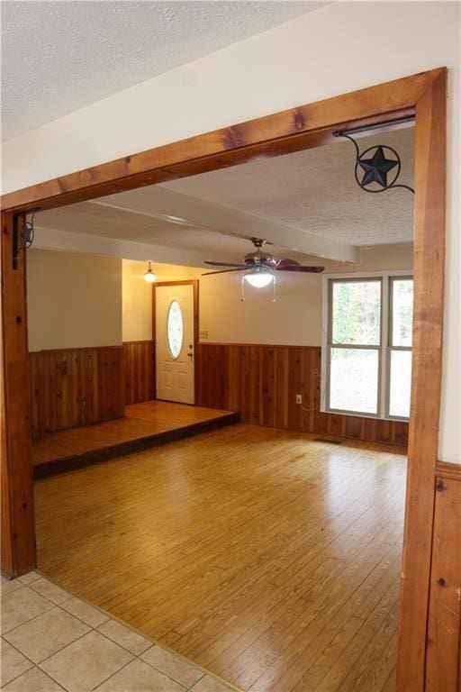 unfurnished living room featuring wainscoting, wood walls, light wood finished floors, ceiling fan, and a textured ceiling