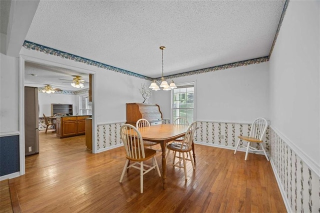 dining area with a textured ceiling, wainscoting, ceiling fan with notable chandelier, wallpapered walls, and hardwood / wood-style flooring