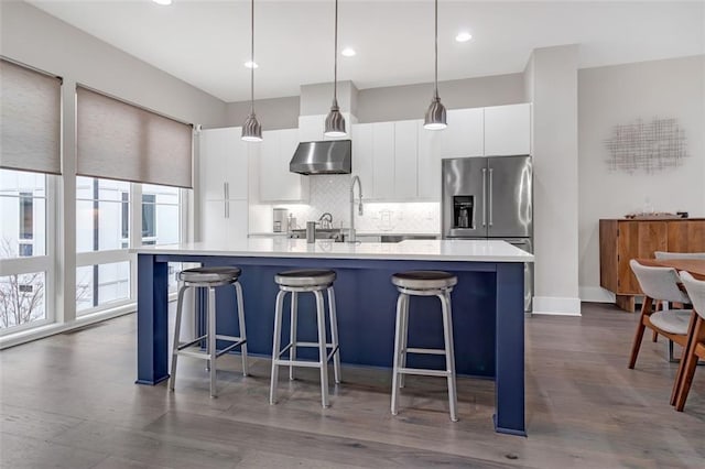 kitchen featuring white cabinets, wall chimney exhaust hood, decorative light fixtures, tasteful backsplash, and a center island with sink