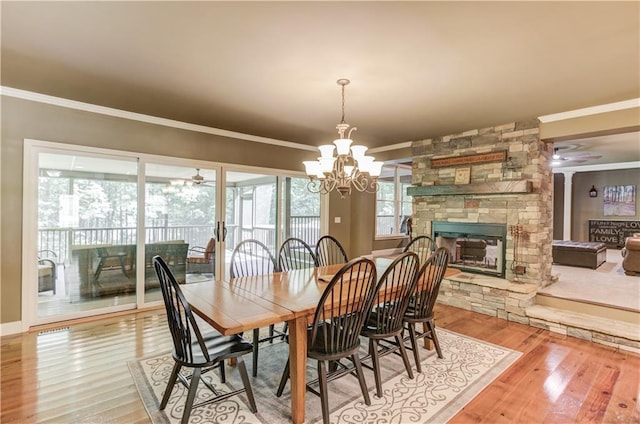 dining area featuring hardwood / wood-style flooring, crown molding, a stone fireplace, and ceiling fan with notable chandelier