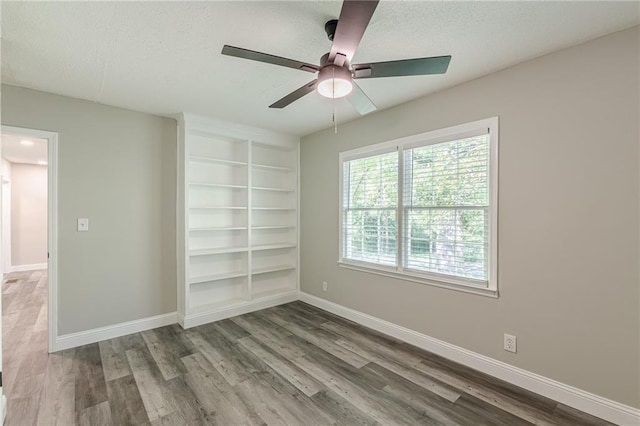 unfurnished room with wood finished floors, a ceiling fan, baseboards, a textured ceiling, and built in shelves