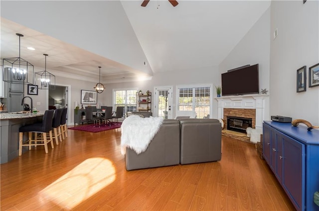 living area with light wood-type flooring, high vaulted ceiling, a ceiling fan, a fireplace, and a raised ceiling