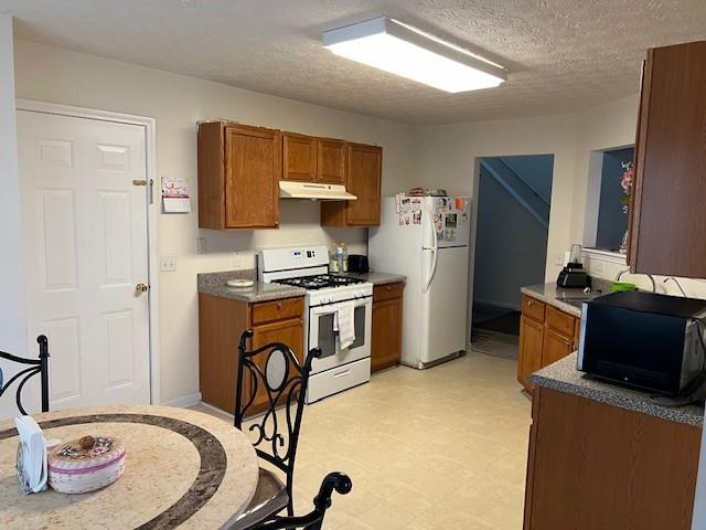 kitchen with brown cabinets, light floors, dark countertops, white appliances, and under cabinet range hood