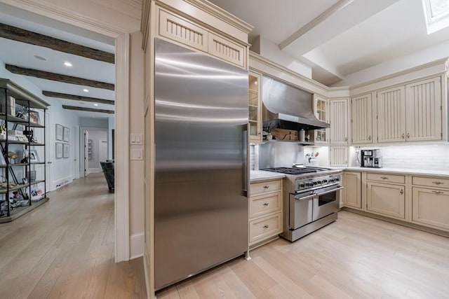 kitchen with high end appliances, cream cabinetry, wall chimney range hood, and beamed ceiling
