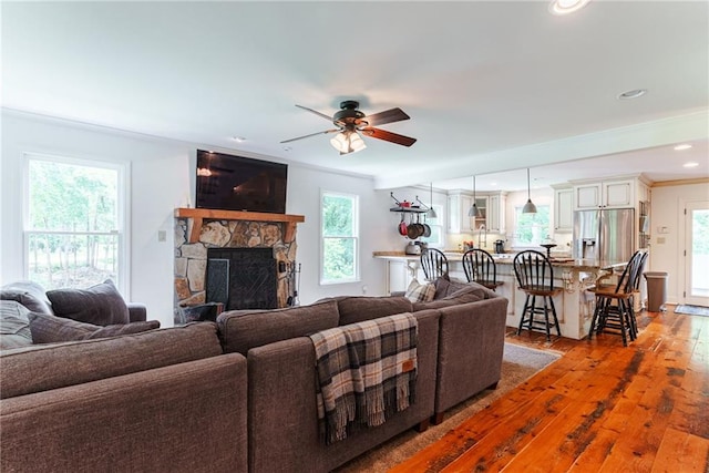 living room with ceiling fan, a stone fireplace, hardwood / wood-style flooring, and crown molding