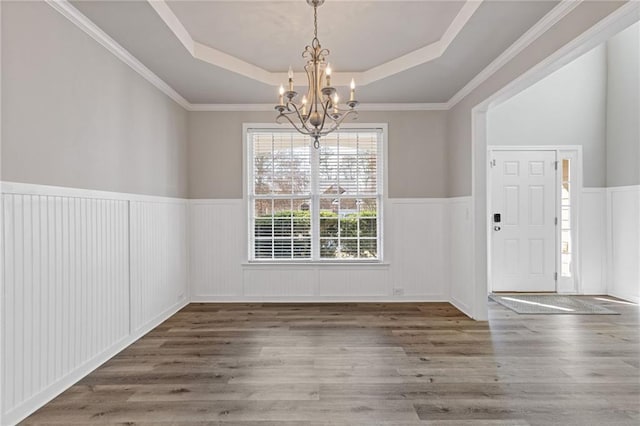 unfurnished dining area with an inviting chandelier, a tray ceiling, wood finished floors, and a wainscoted wall