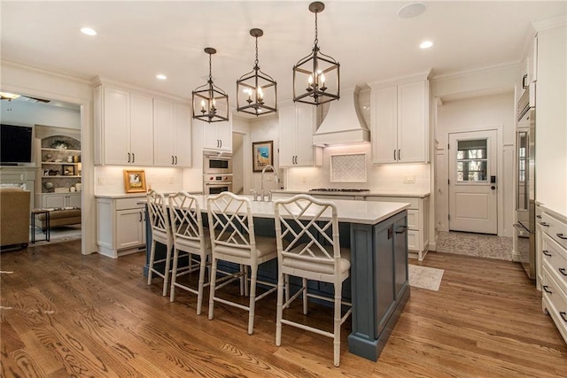 kitchen featuring custom range hood, decorative light fixtures, hardwood / wood-style flooring, a center island with sink, and white cabinetry