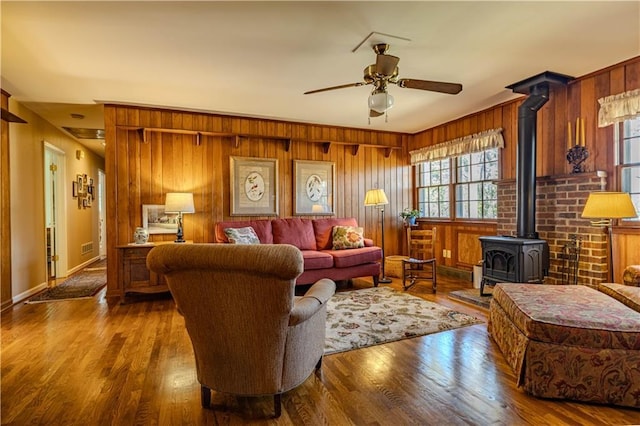 living area featuring a ceiling fan, a wood stove, wood walls, wood finished floors, and baseboards