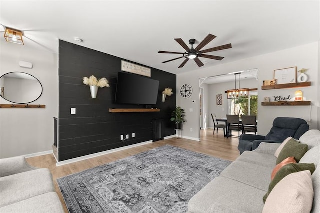 living area featuring an accent wall, ceiling fan with notable chandelier, baseboards, and wood finished floors