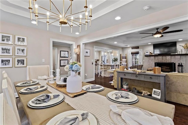 dining space featuring a raised ceiling, a fireplace, ceiling fan with notable chandelier, and dark wood-type flooring