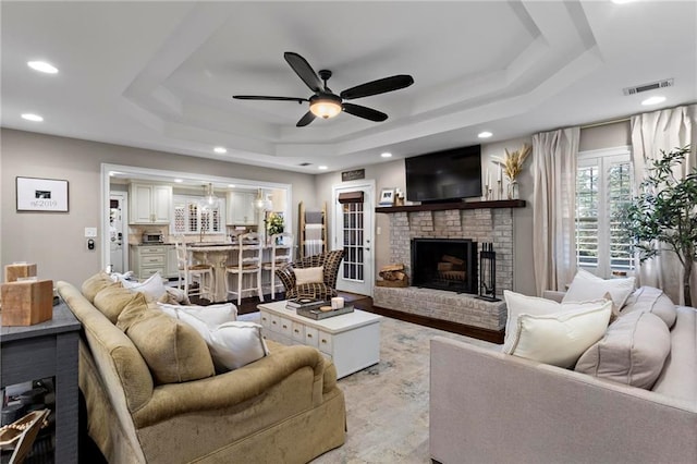 living room featuring a brick fireplace, a tray ceiling, and ceiling fan