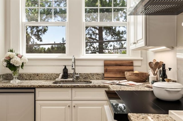 kitchen with a sink, light stone counters, stainless steel dishwasher, white cabinets, and wall chimney range hood