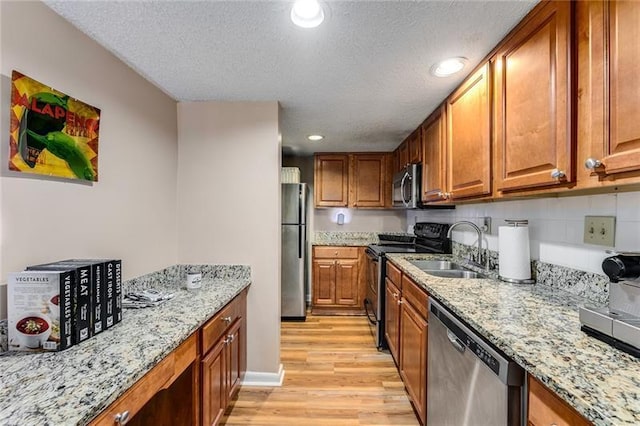 kitchen with a sink, brown cabinetry, light wood-style flooring, light stone counters, and appliances with stainless steel finishes