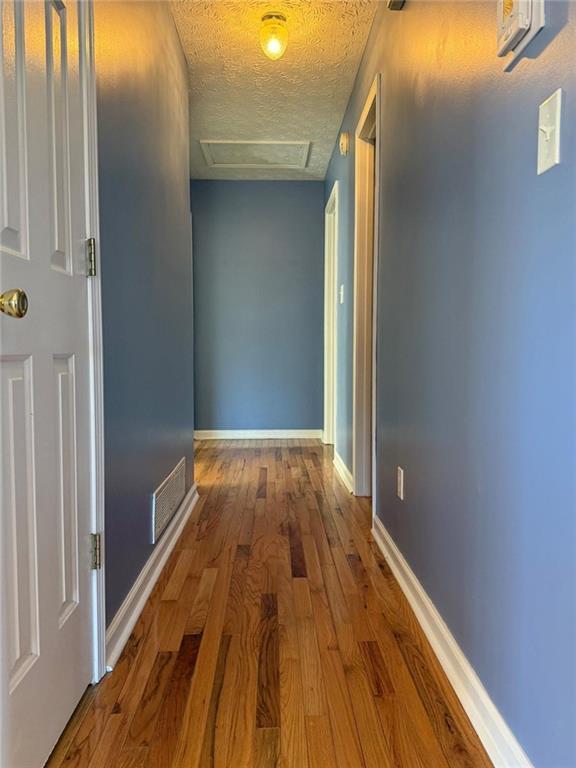 hallway with a textured ceiling, wood finished floors, visible vents, baseboards, and attic access