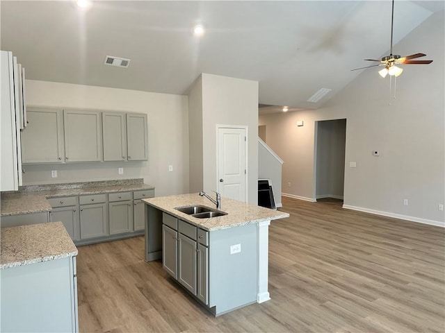 kitchen with an island with sink, light stone countertops, vaulted ceiling, light wood-type flooring, and a sink