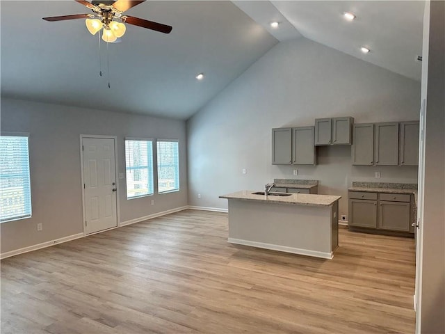 kitchen featuring baseboards, an island with sink, light wood-style flooring, light stone counters, and a sink