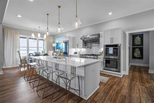 kitchen with wall chimney range hood, decorative light fixtures, stainless steel appliances, and a center island with sink