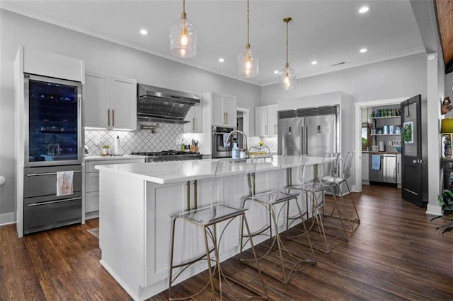 kitchen with wall chimney exhaust hood, a breakfast bar, hanging light fixtures, a large island with sink, and white cabinets