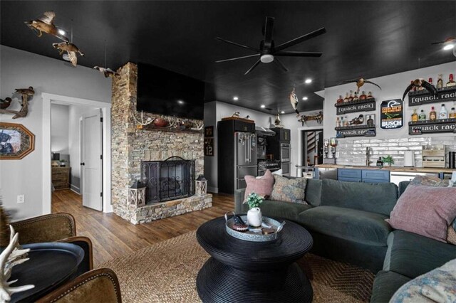 living room featuring hardwood / wood-style flooring, a stone fireplace, indoor wet bar, and ceiling fan