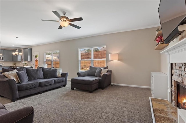 carpeted living room featuring a stone fireplace, ceiling fan, baseboards, a chandelier, and ornamental molding