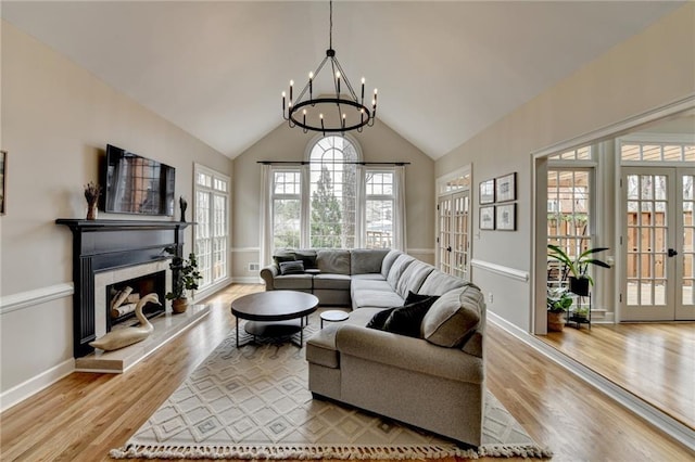 living room with french doors, a chandelier, a fireplace with raised hearth, light wood-style floors, and baseboards