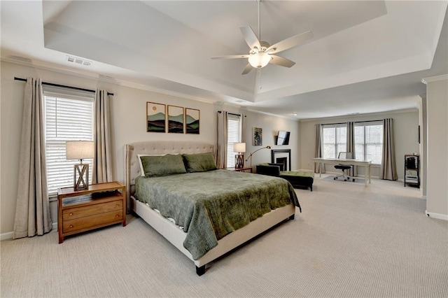 carpeted bedroom with a tray ceiling, ornamental molding, baseboards, a fireplace, and a ceiling fan