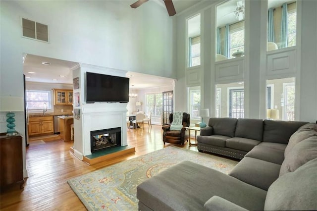 living area featuring a high ceiling, a ceiling fan, light wood-style flooring, and a glass covered fireplace