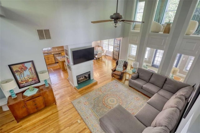 living area with light wood-type flooring, plenty of natural light, a high ceiling, a ceiling fan, and a fireplace with raised hearth
