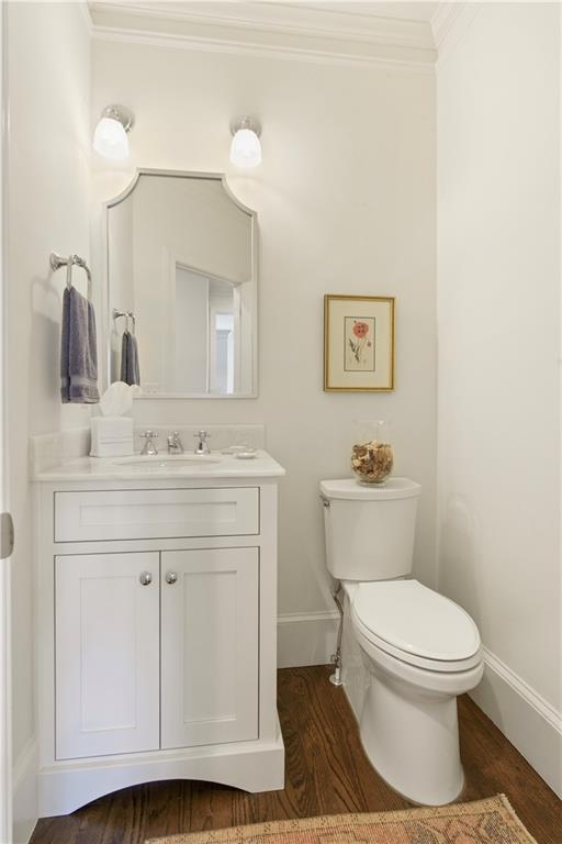 half bath with toilet, wood finished floors, vanity, baseboards, and crown molding