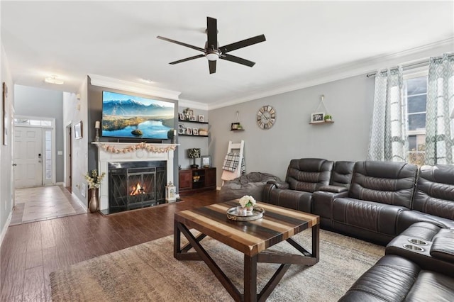 living area with ornamental molding, ceiling fan, wood finished floors, a warm lit fireplace, and baseboards