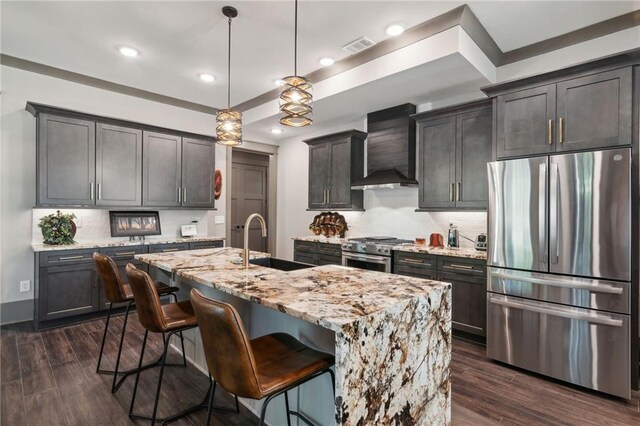 kitchen with decorative light fixtures, a center island with sink, wall chimney exhaust hood, stainless steel appliances, and dark hardwood / wood-style flooring