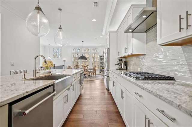 kitchen featuring a sink, backsplash, appliances with stainless steel finishes, wall chimney range hood, and dark wood-style flooring