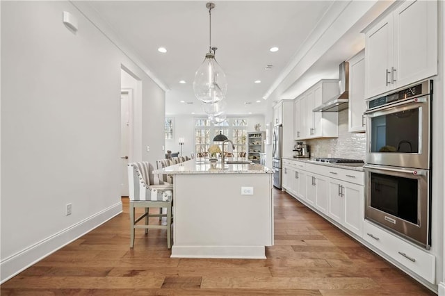 kitchen with light wood-style flooring, a kitchen island with sink, stainless steel appliances, wall chimney exhaust hood, and backsplash