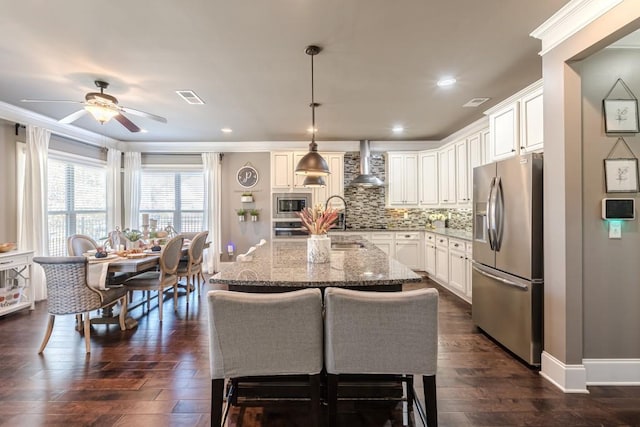 kitchen featuring a kitchen island with sink, wall chimney range hood, hanging light fixtures, light stone countertops, and appliances with stainless steel finishes