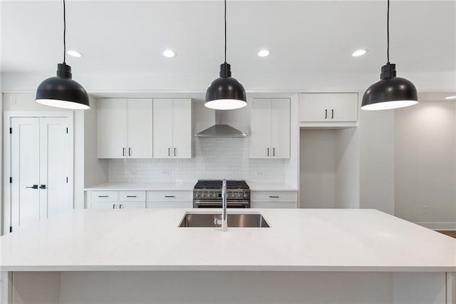 kitchen with wall chimney range hood, white cabinets, hanging light fixtures, and a kitchen island with sink